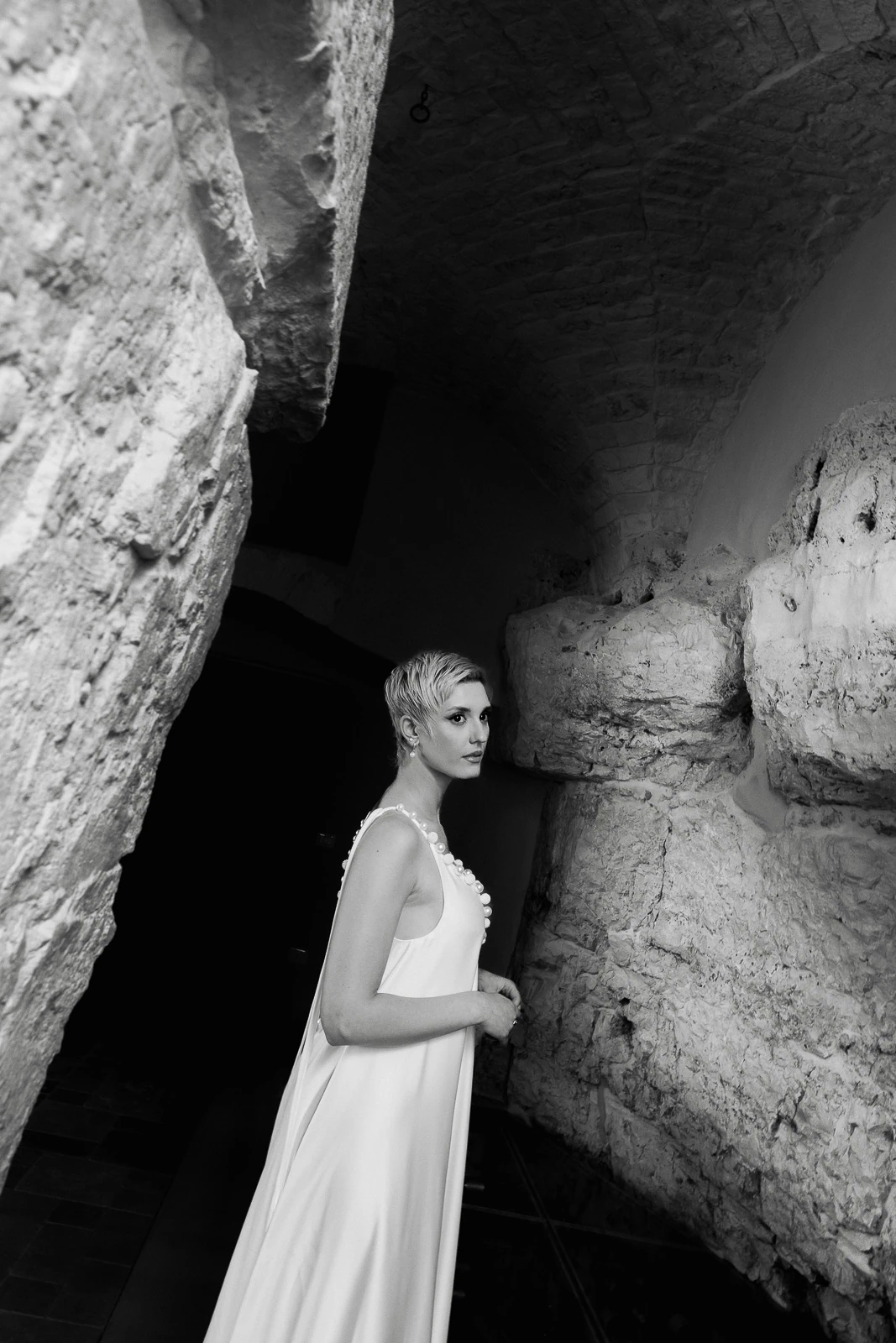 A bride in an elegant, sleeveless gown stands beside a historic stone wall in a softly lit Puglia, Italy venue.