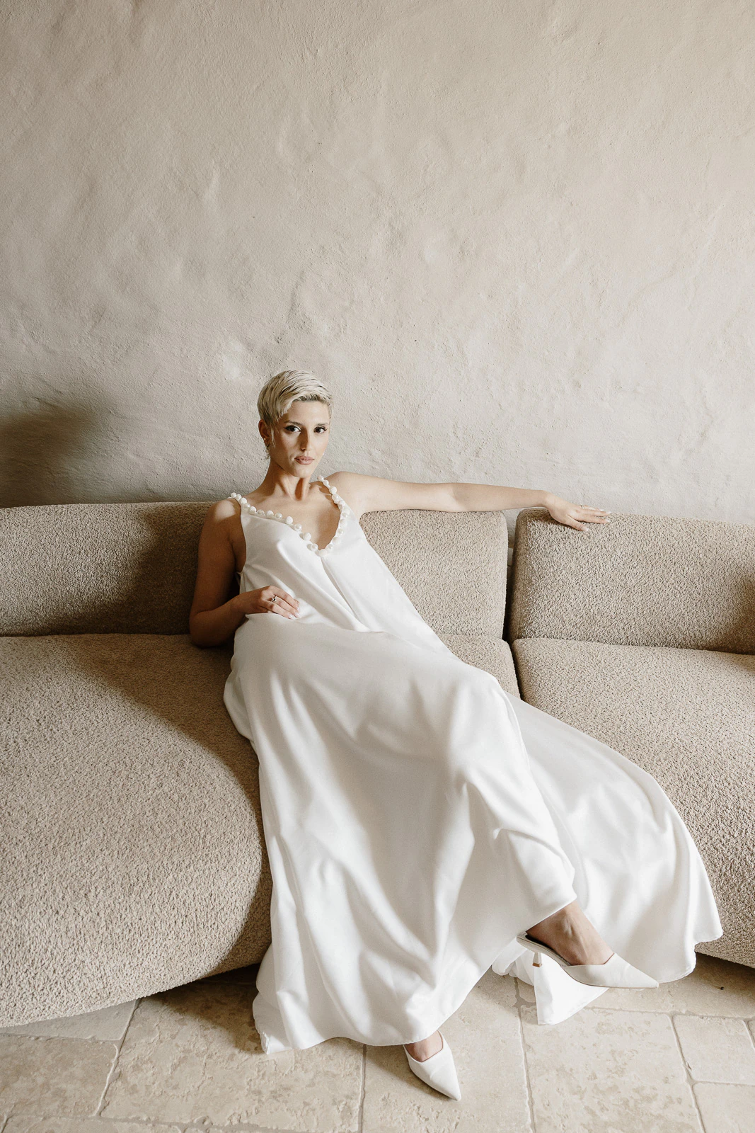 A bride with short blonde hair in an elegant white gown sits gracefully on a textured sofa, exuding timeless Puglia romance.