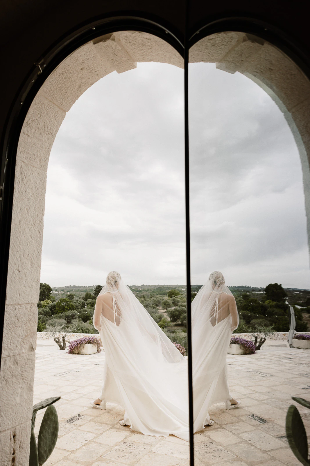 An elegant bride in Puglia stands by a stone archway, her reflection shimmering in glass against the Italian countryside.