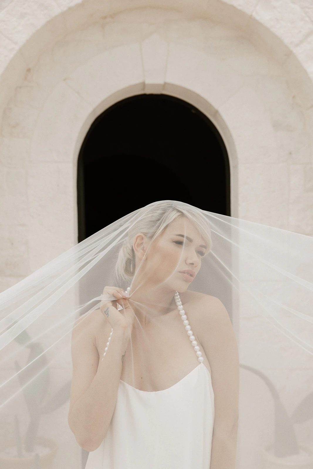 A bride gracefully lifts her veil beneath a centuries-old arched doorway in Puglia, exuding elegance and anticipation.