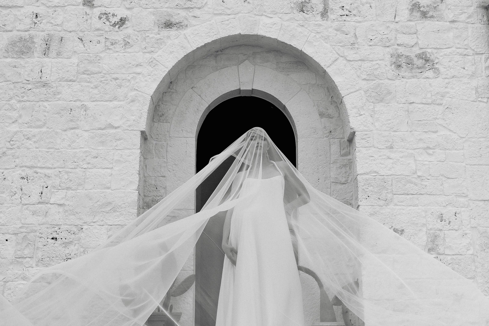 A bride in a flowing veil lingers on sunlit stone steps before an arched doorway, evoking timeless romance in Puglia.
