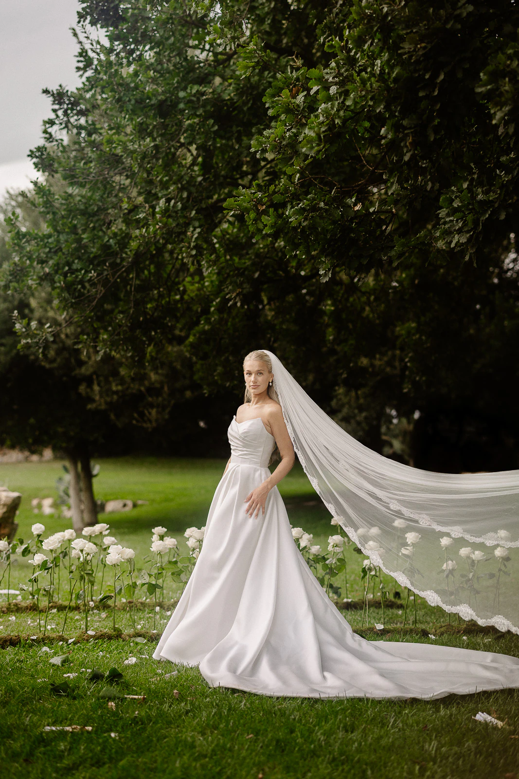 A bride in a flowing white gown stands amid blooming white flowers and olive trees, capturing romance in sunlit Puglia.