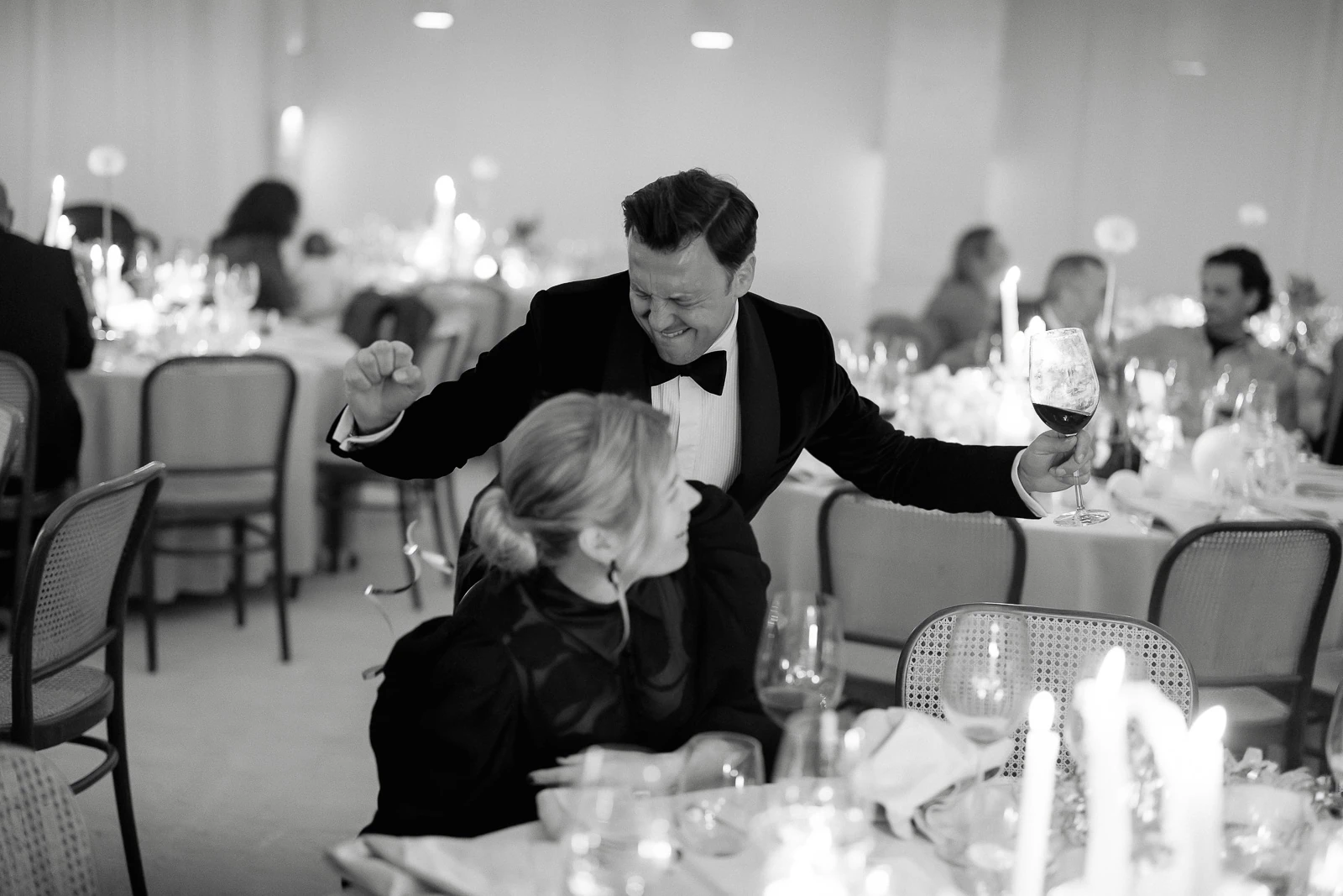 At an elegant candlelit celebration in Puglia, a tuxedoed guest toasts joyfully with a seated companion amid lively conversation.