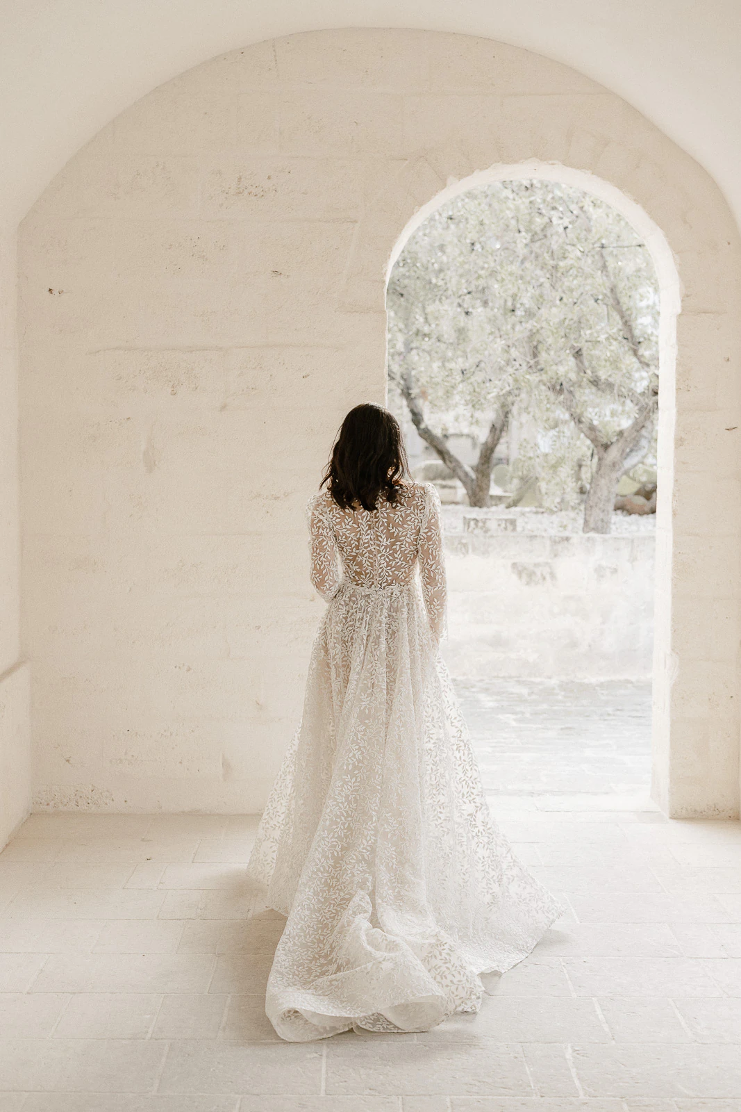 A bride in a flowing white lace gown gazes through an arched doorway in Puglia, sunlight illuminating Italian olive trees.