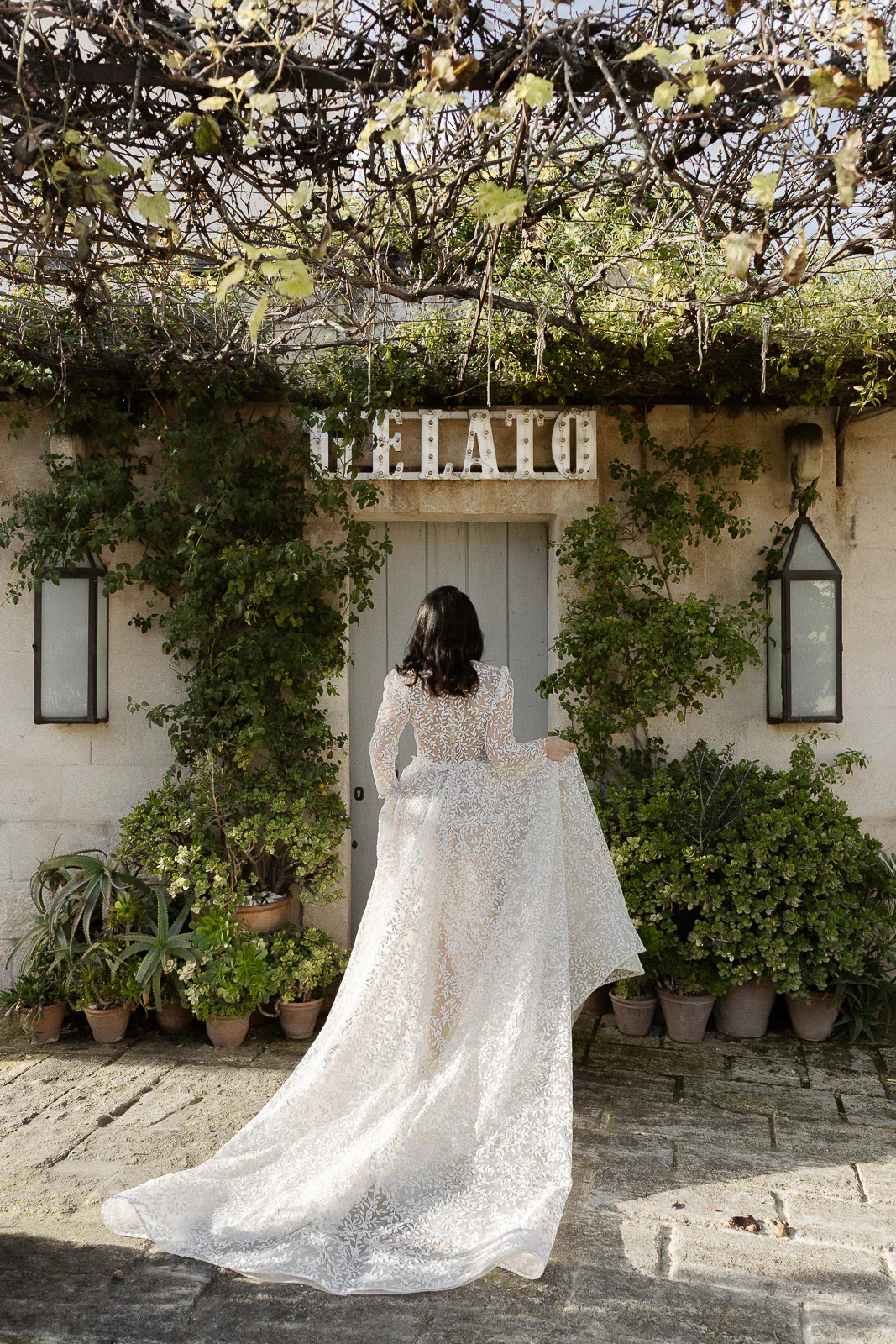 A bride in elegant lace gazes toward a vine-draped doorway, surrounded by lush greenery in a rustic Puglia setting.