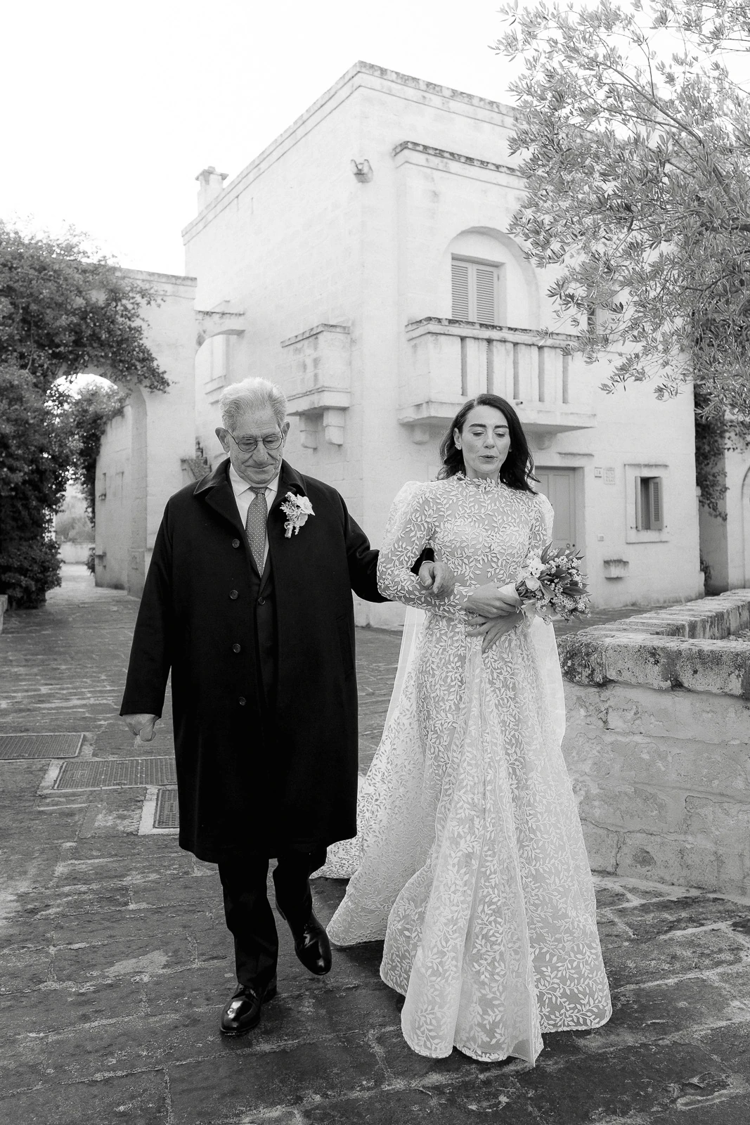 A radiant bride in lace strolls arm in arm with her father through sunlit stone arches in romantic Puglia, Italy.