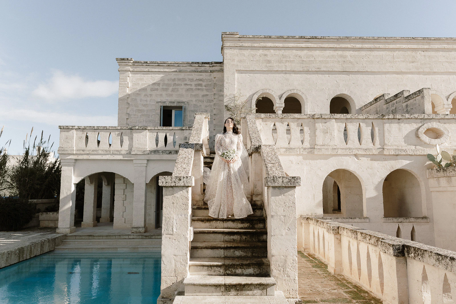 A bride ascends sunlit stone steps at a grand Puglia villa, serene pool glistening beneath arched Italian windows.