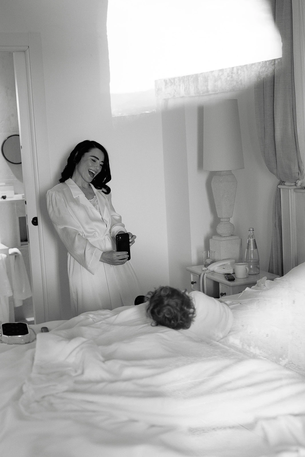 In sunlit Puglia, a mother in a robe captures a tender morning moment with her child nestled beneath soft linens.