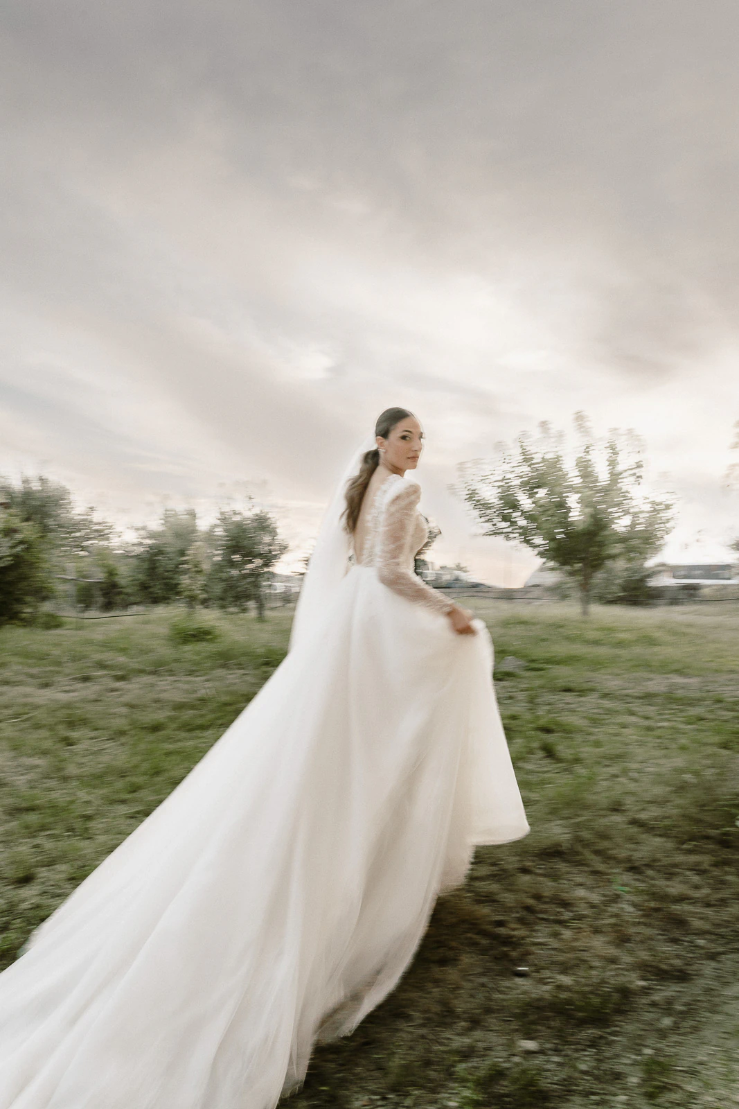 A bride glances back gracefully as she strolls across lush grass beneath dramatic skies in romantic Puglia, Italy.