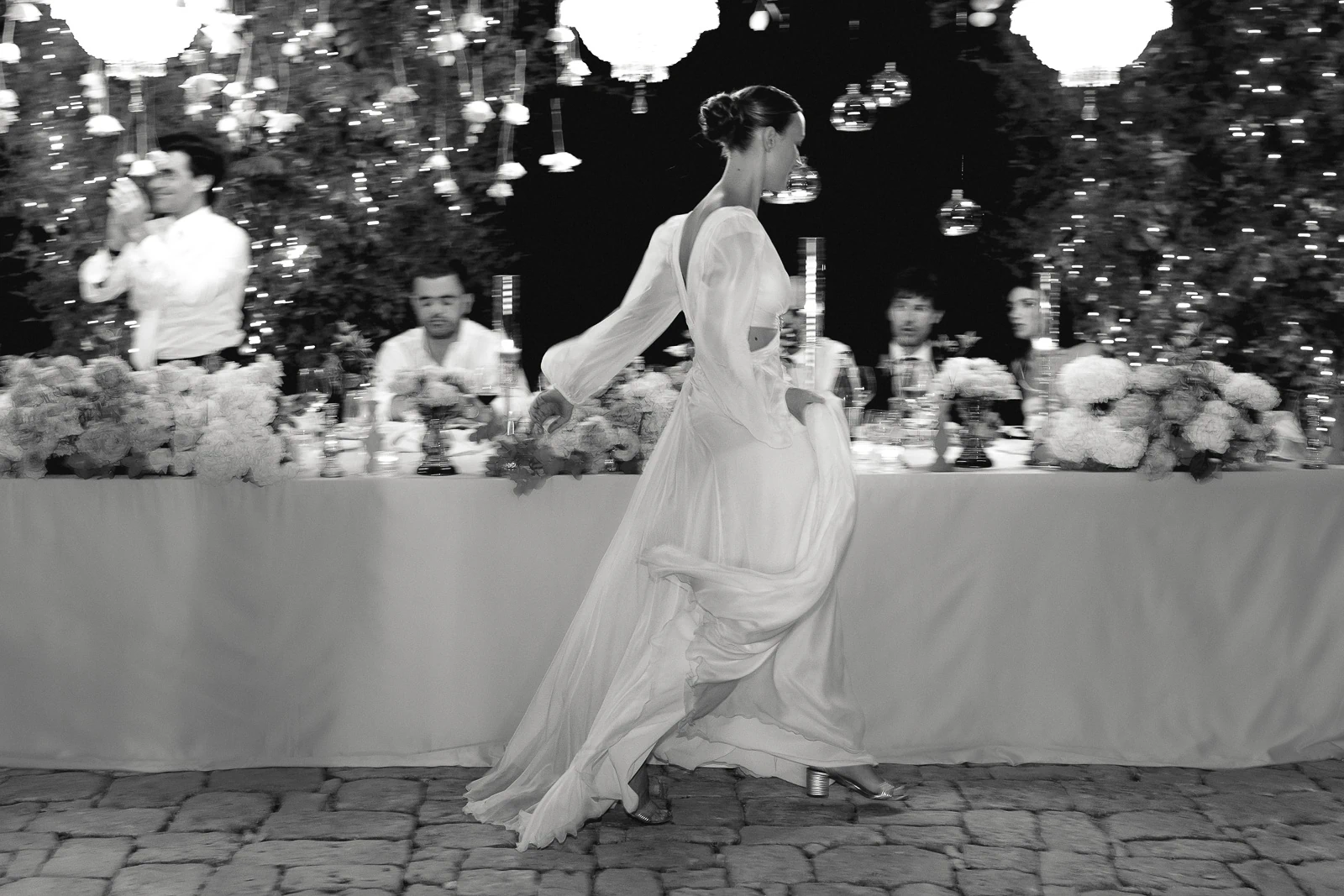 In Puglia, a woman in an elegant gown glides past guests beneath cascading florals and glowing lights at a romantic wedding table.