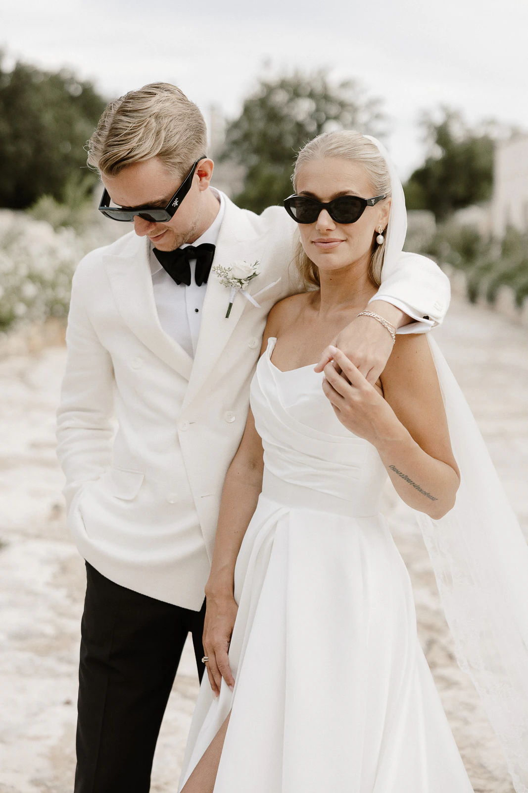 Chic bride and groom in sunglasses share a sunlit moment amid Puglia’s romantic landscape, embracing la dolce vita.