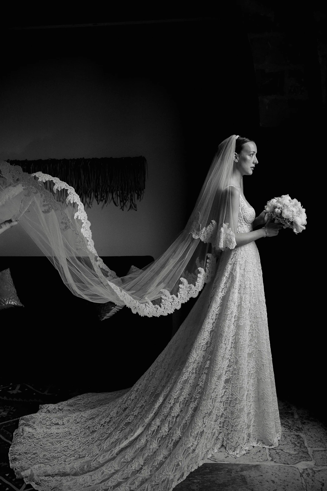 In an atmospheric Italian villa, a bride in lace and a sweeping veil stands aglow, bouquet in hand beneath moody lighting.