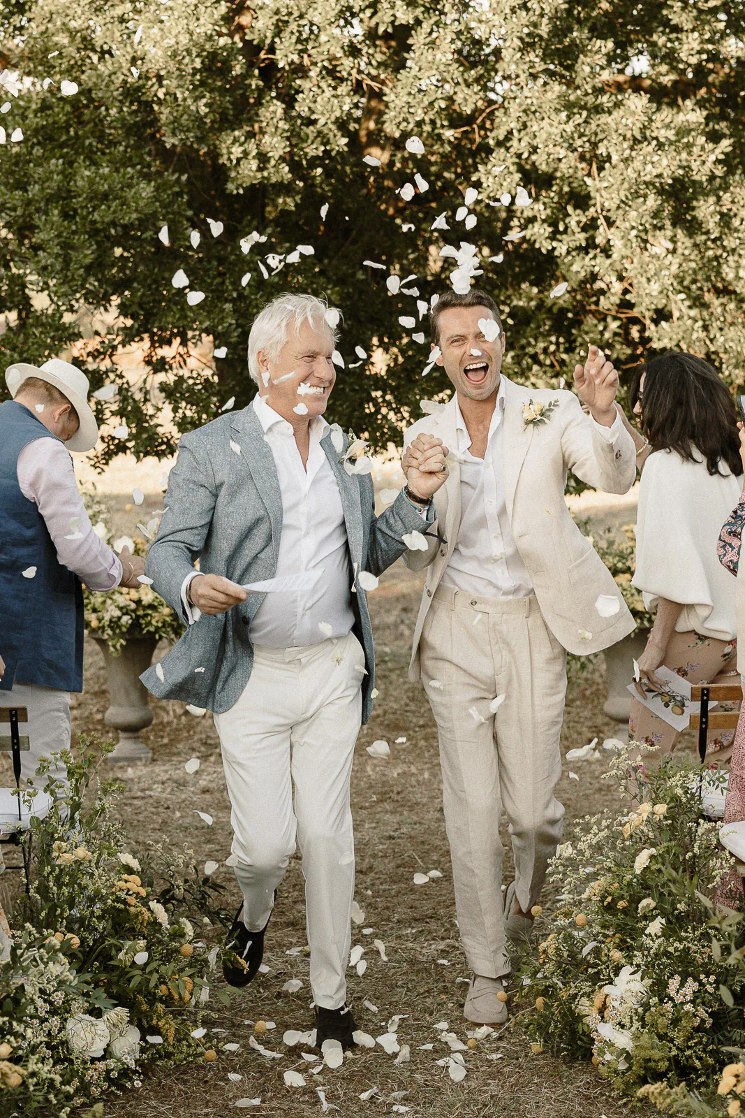 Newlyweds in light suits walk hand in hand beneath a grand Puglian tree, flower petals swirling in celebration.