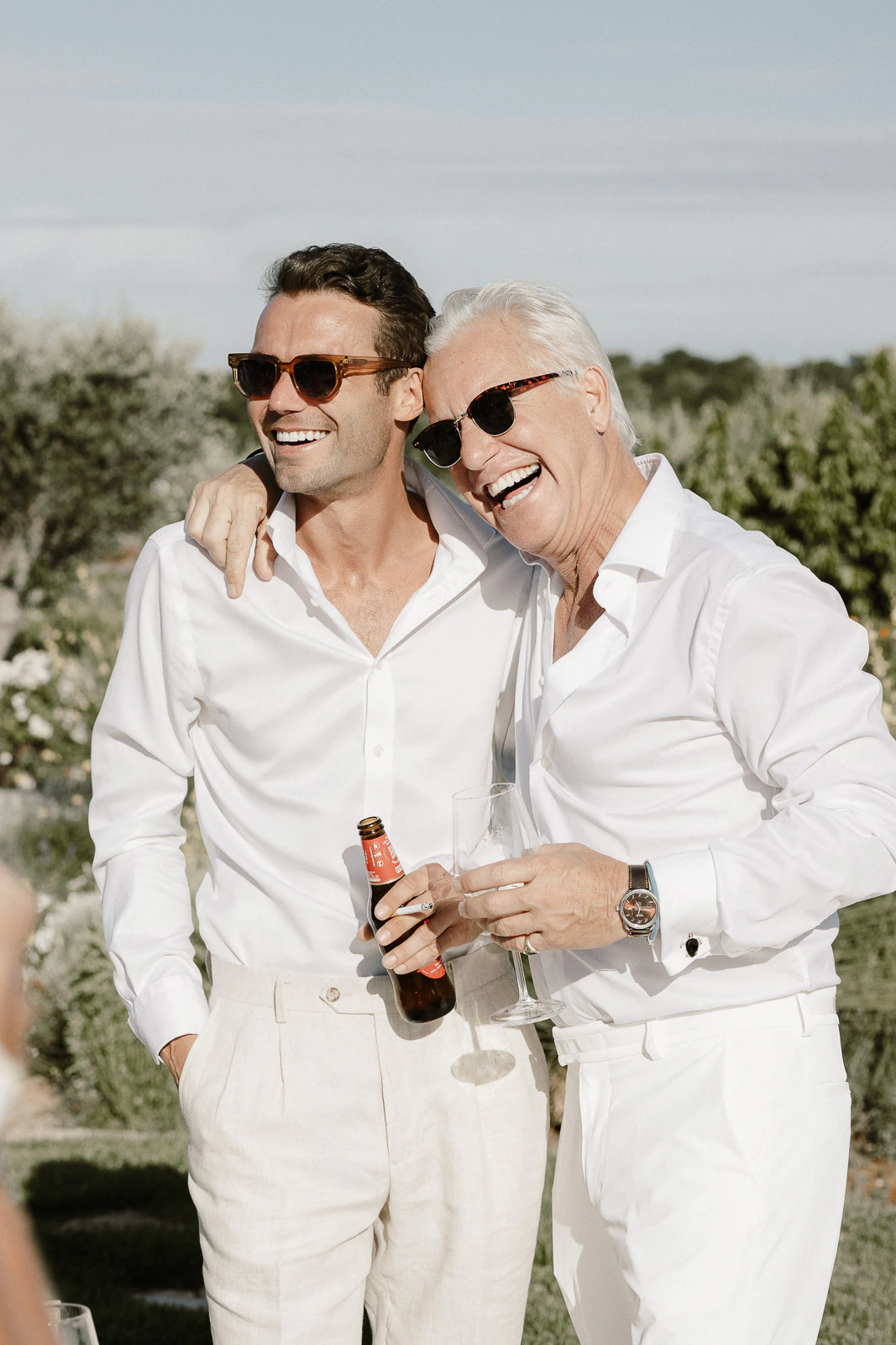 Amid sunlit greenery in Puglia, two men in crisp white shirts share laughter and drinks, capturing Italian wedding joy.