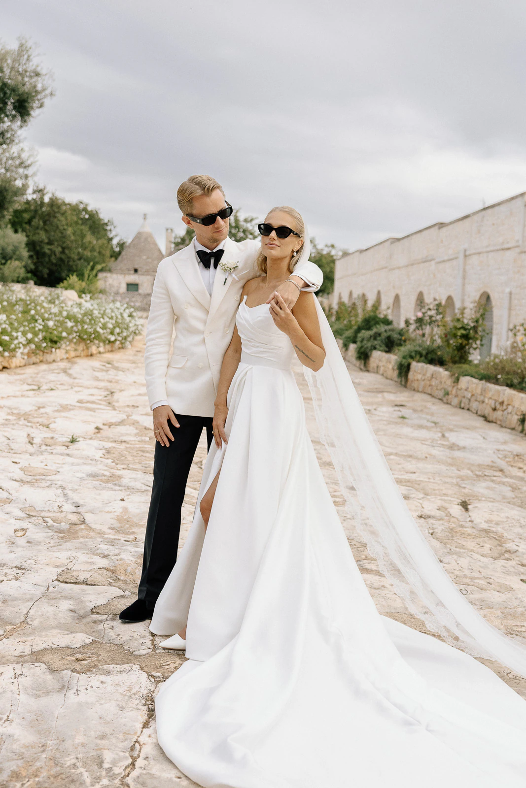Stylish newlyweds in sunglasses share a joyful moment on a sunlit stone path in romantic Puglia, Italy.