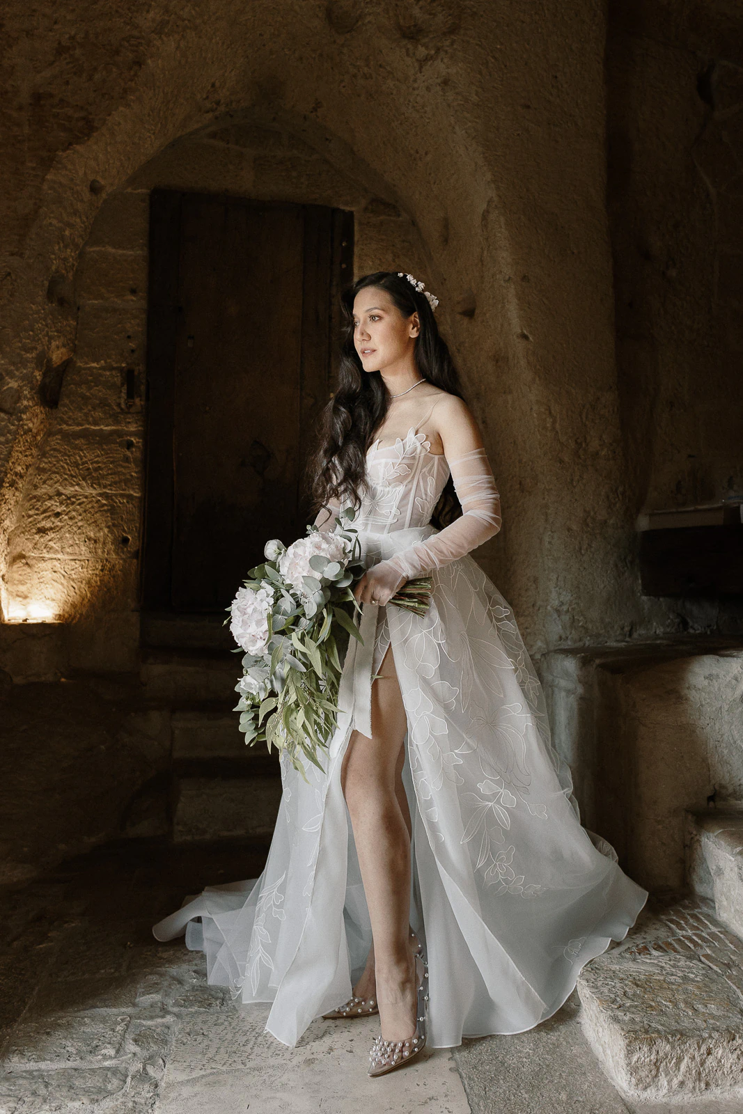A bride in a floral off-shoulder gown stands by a rustic stone doorway in Puglia, softly holding white blooms.