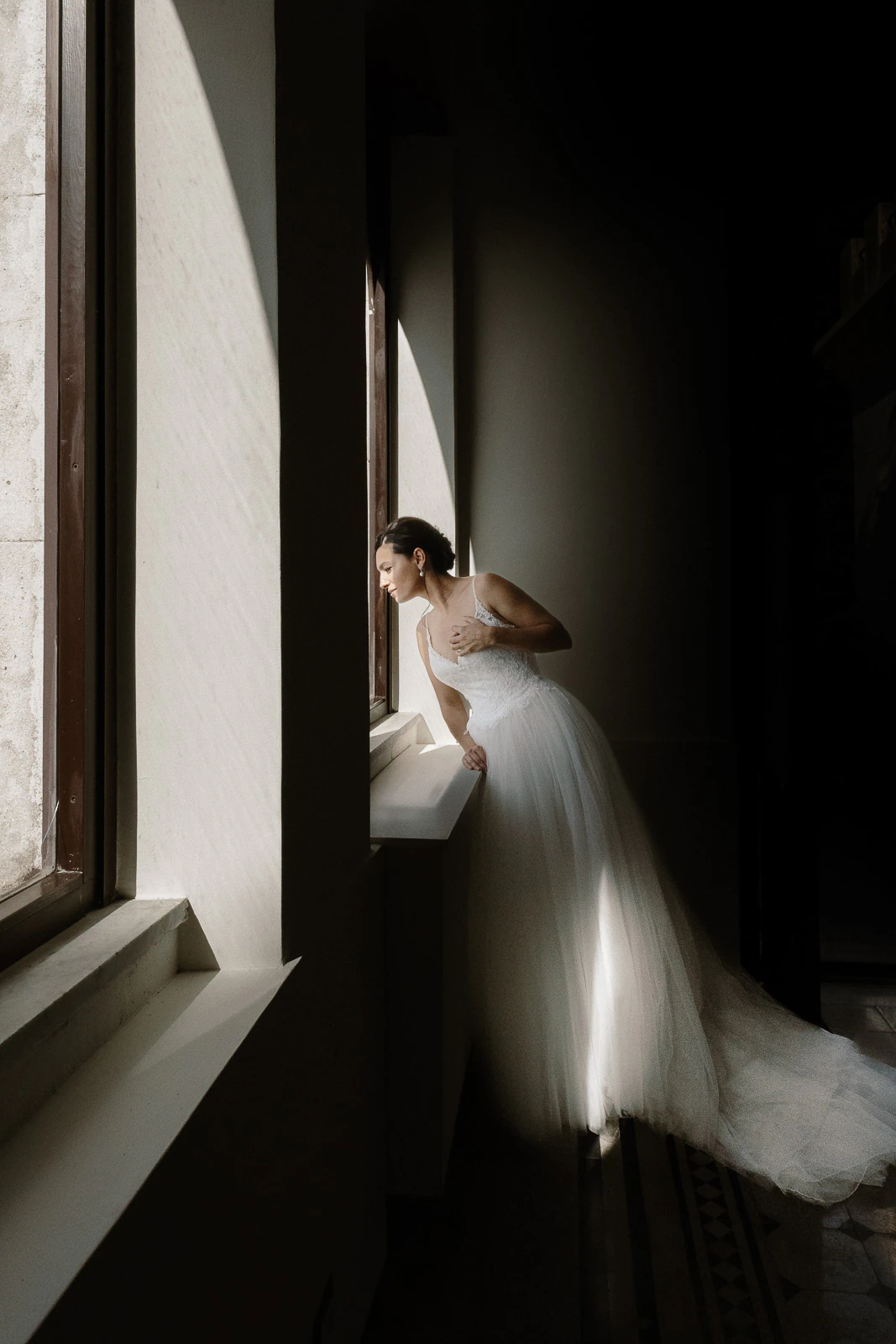 A bride in a luminous white gown gazes out a sunlit Puglia window, soft shadows casting an intimate, timeless ambiance.