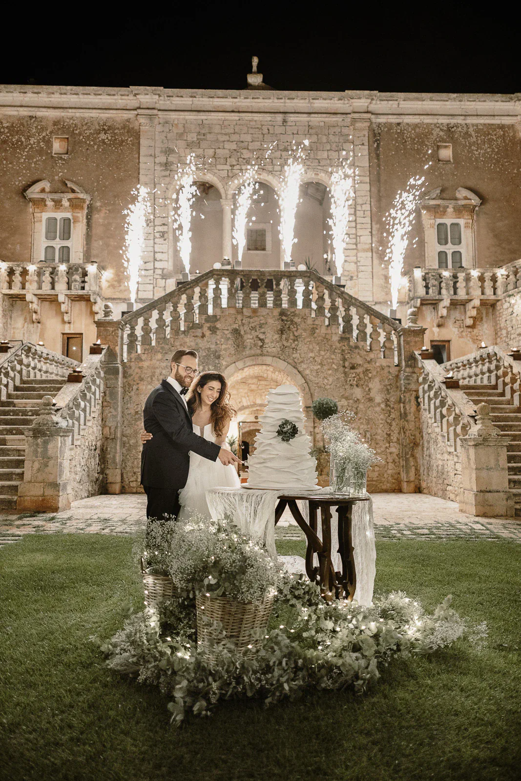 Under a starlit Puglian sky, a couple cuts their wedding cake as fireworks illuminate an elegant Italian villa.