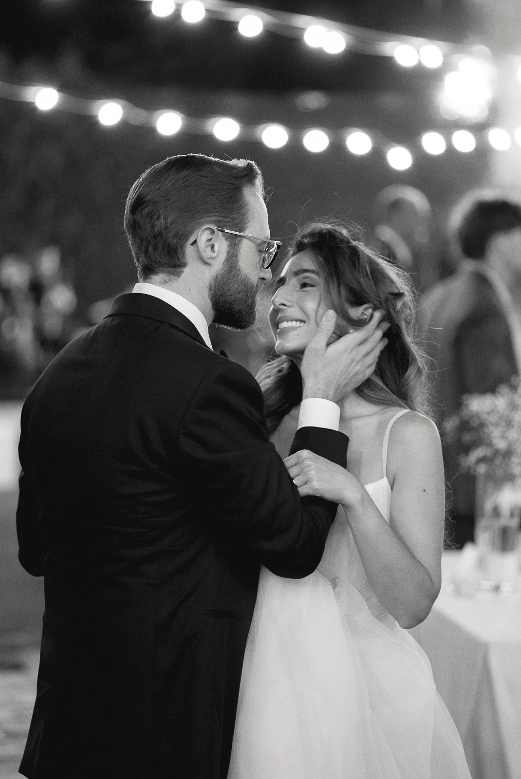 The bride and groom share an intimate dance beneath glowing string lights at their elegant Puglia wedding celebration.
