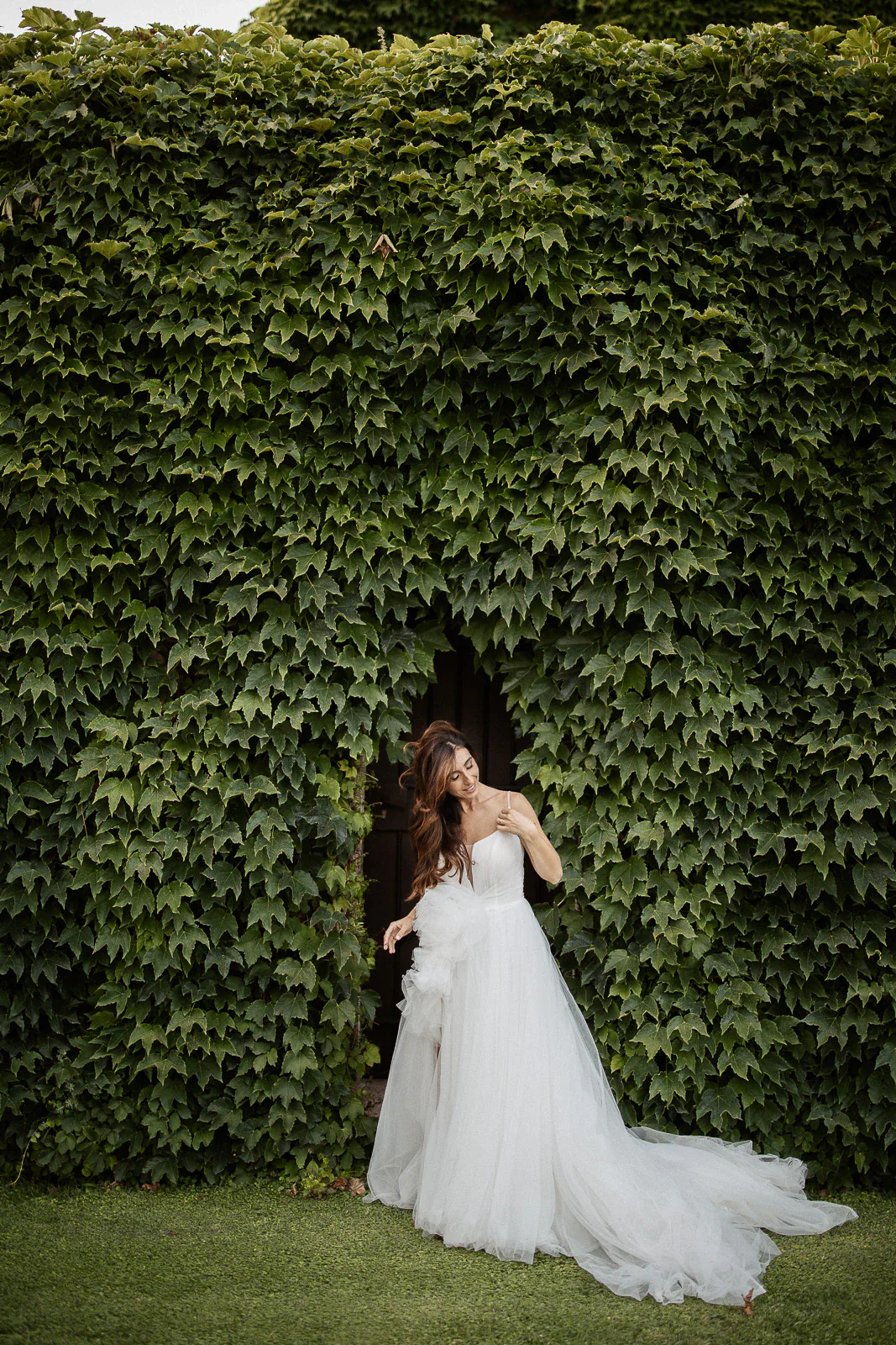 A bride in an ethereal white gown stands amidst lush ivy in Puglia, Italy, gazing upward in quiet anticipation.