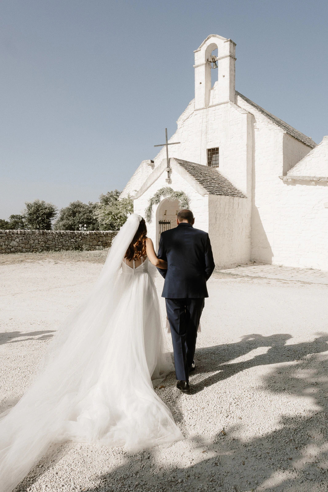In sunlit Puglia, a bride and her escort approach a white stone church, capturing timeless romance and Italian elegance.