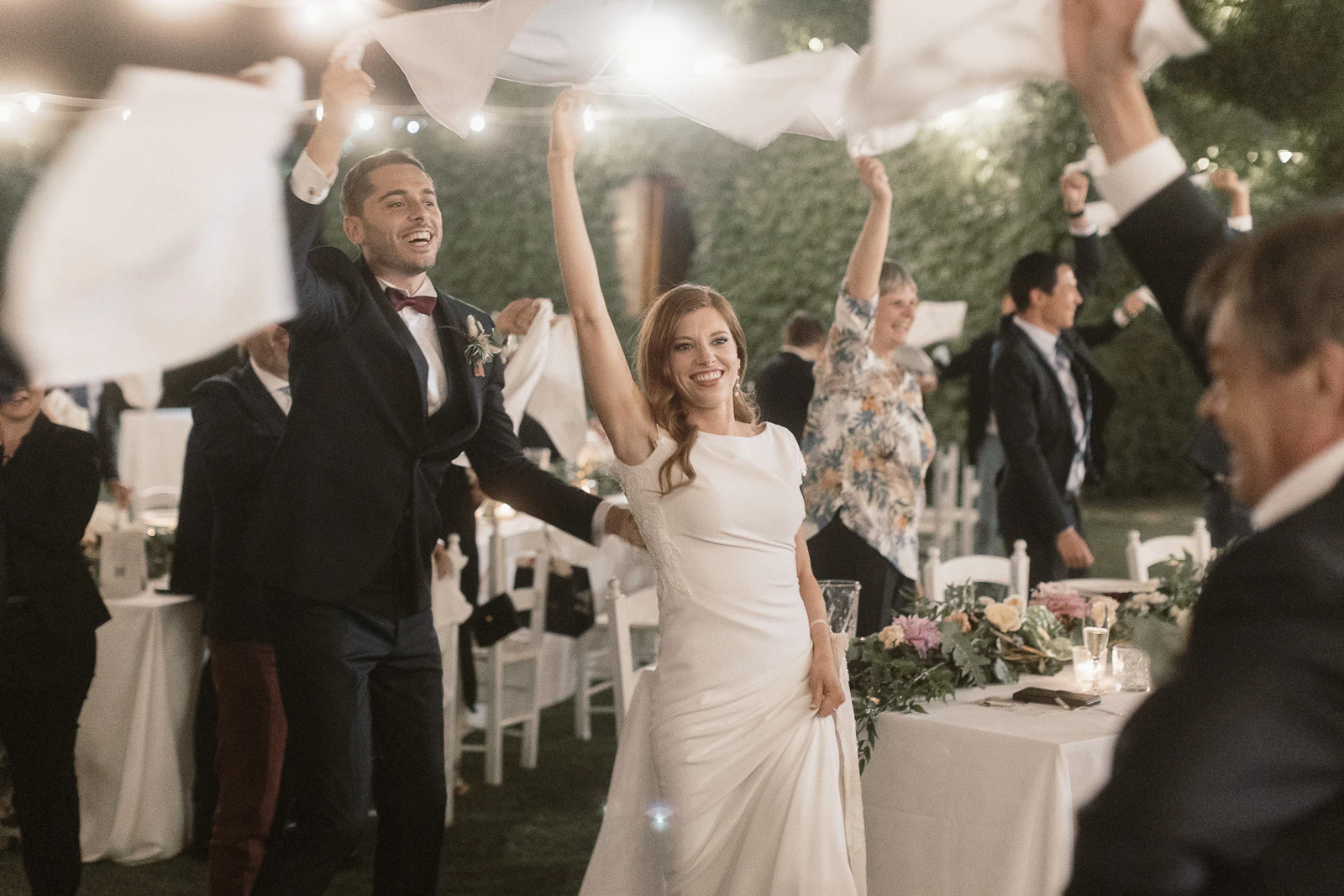In Puglia, a radiant couple enters their reception, guests joyfully waving napkins amid laughter and elegantly adorned tables.