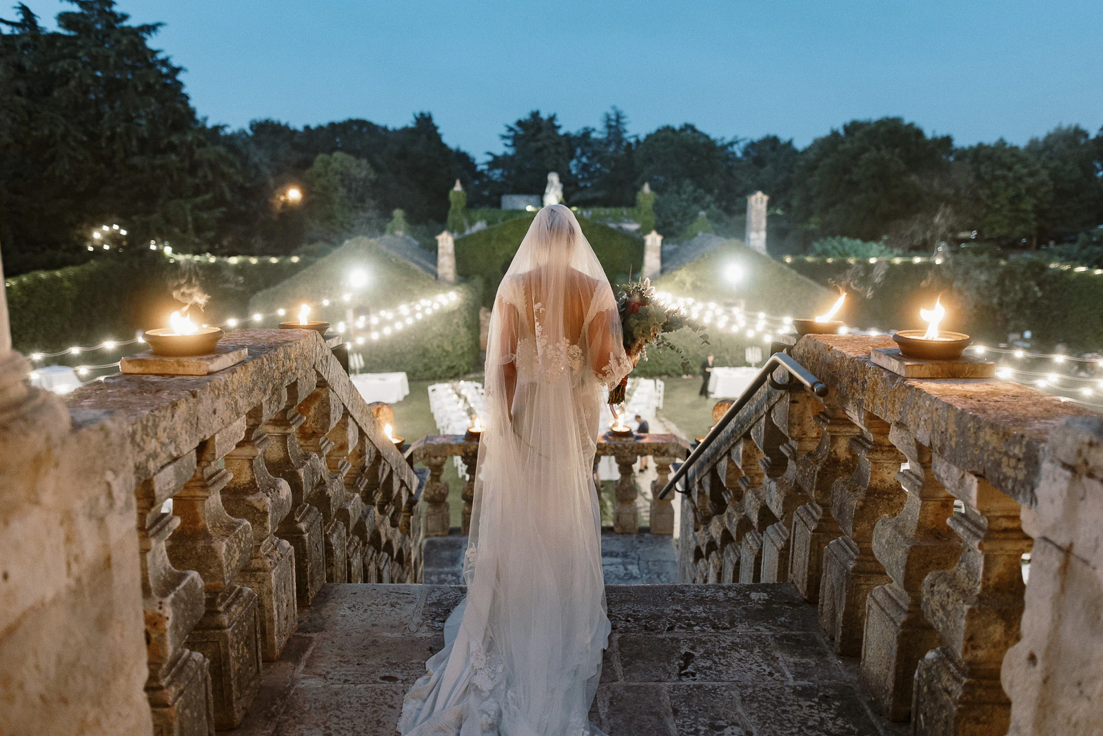 A bride in an elegant white gown stands atop sunlit Puglian steps, framed by glowing lights and lush Mediterranean greenery.