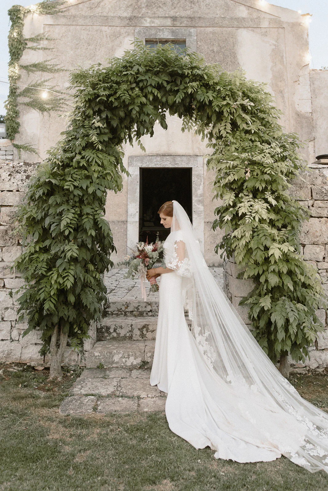 A bride in a flowing white gown stands beneath a lush archway, bouquet in hand, outside a historic Puglia villa.