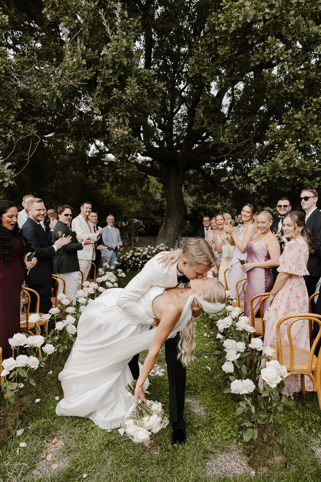 Under a grand tree in Puglia, the groom dips his bride for a romantic kiss amid joyful guests and white roses.