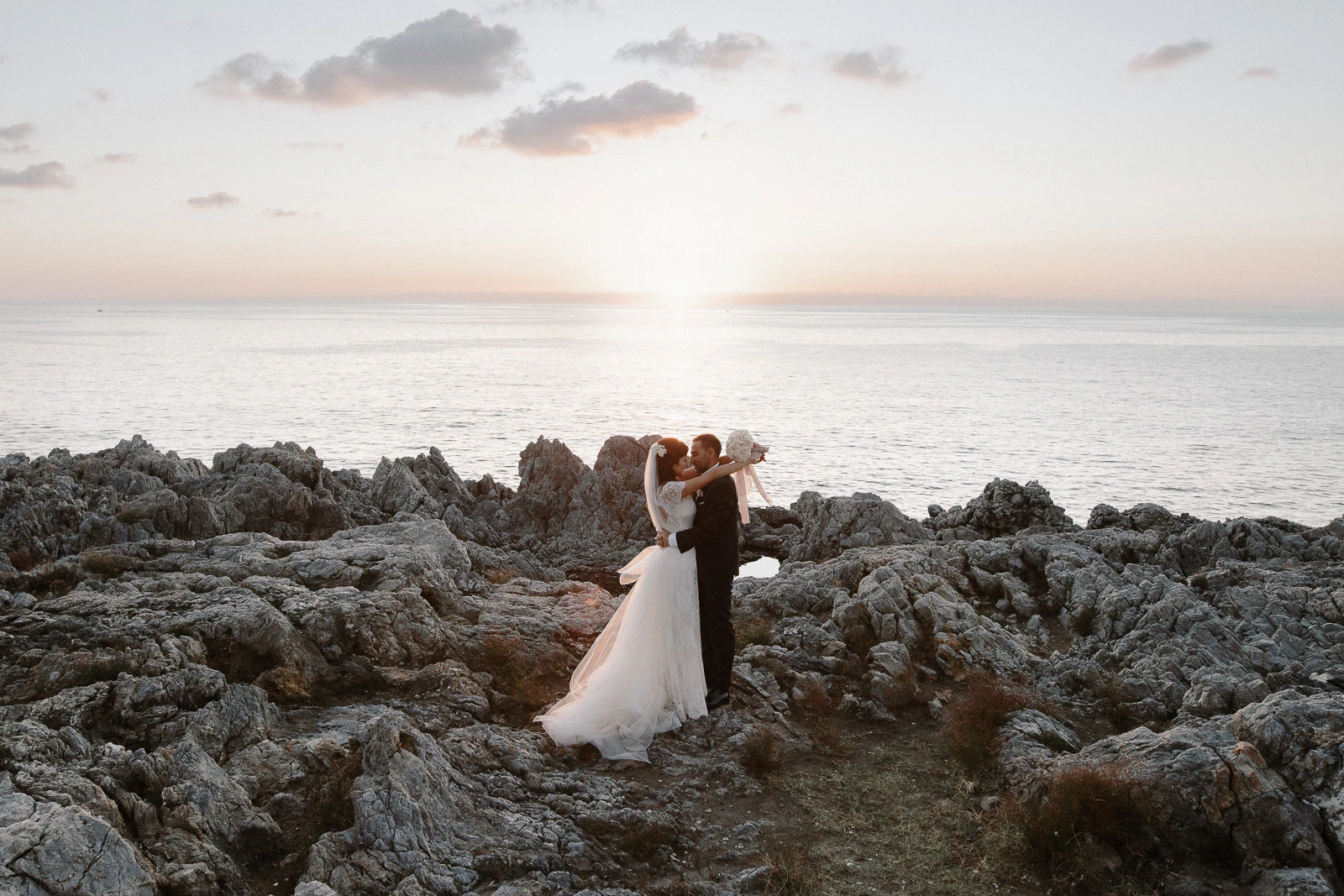 Newlyweds embrace on Puglia’s rocky shoreline at sunset, golden light dancing on the Adriatic and clouds framing their moment.