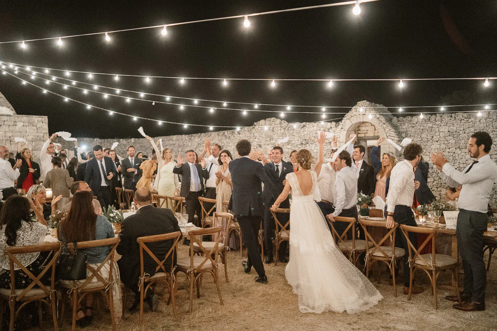 Newlyweds stroll through delighted guests beneath glowing string lights at a joyful Puglia evening wedding celebration.