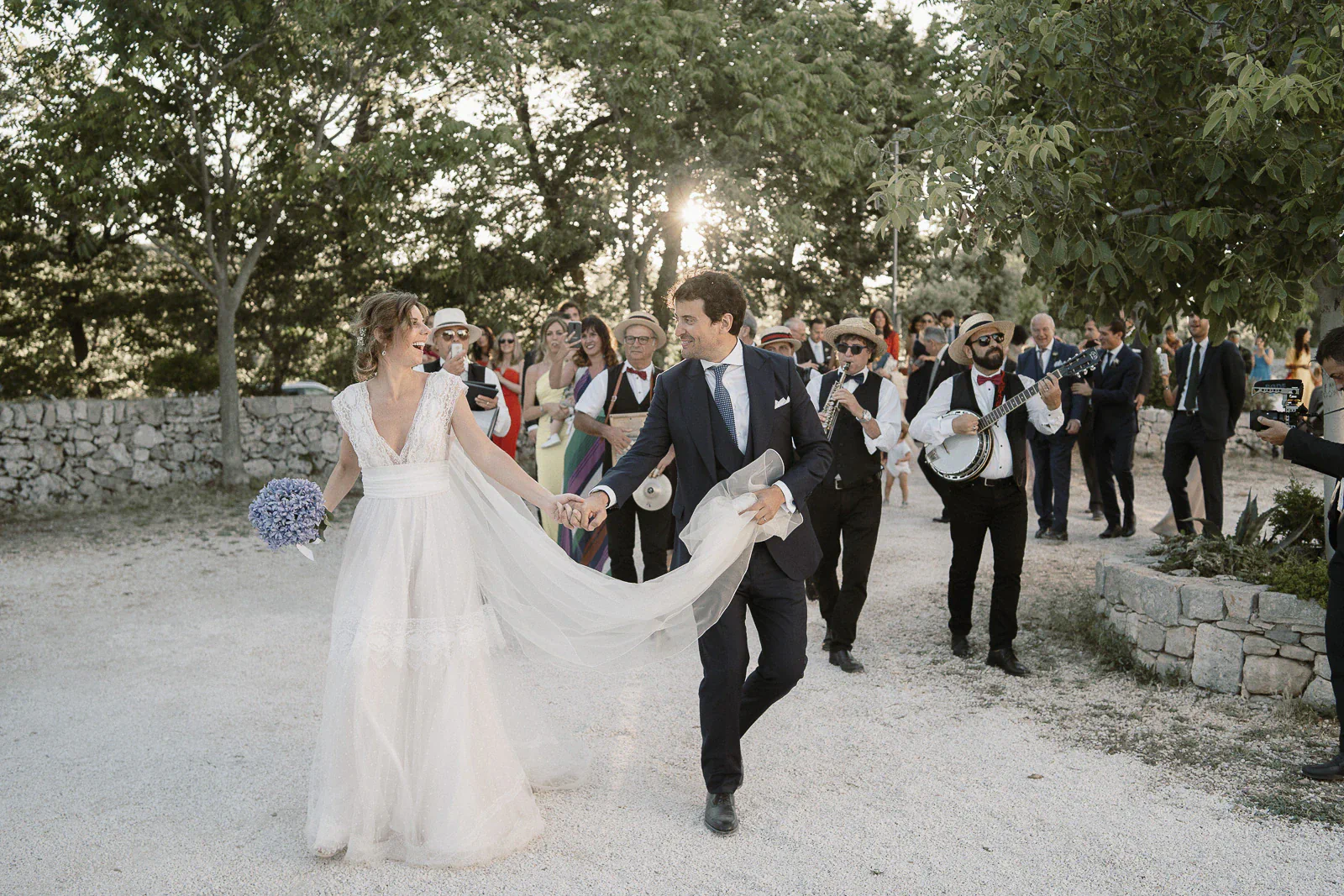 A joyful bride and groom stroll hand in hand through a sunlit Puglian garden, serenaded by musicians and surrounded by loved ones.