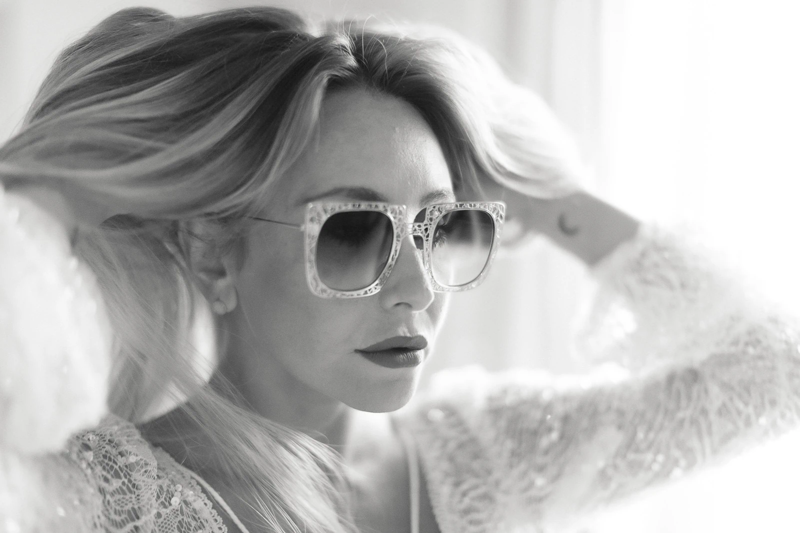 In Puglia, a bride in a delicate lace top adjusts her hair, sunlight streaming through in elegant monochrome allure.