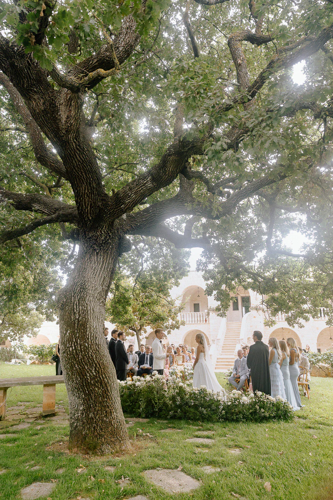 An elegant outdoor ceremony unfolds beneath a grand tree in Puglia, as the couple exchanges vows amid loved ones and sunlit stone.