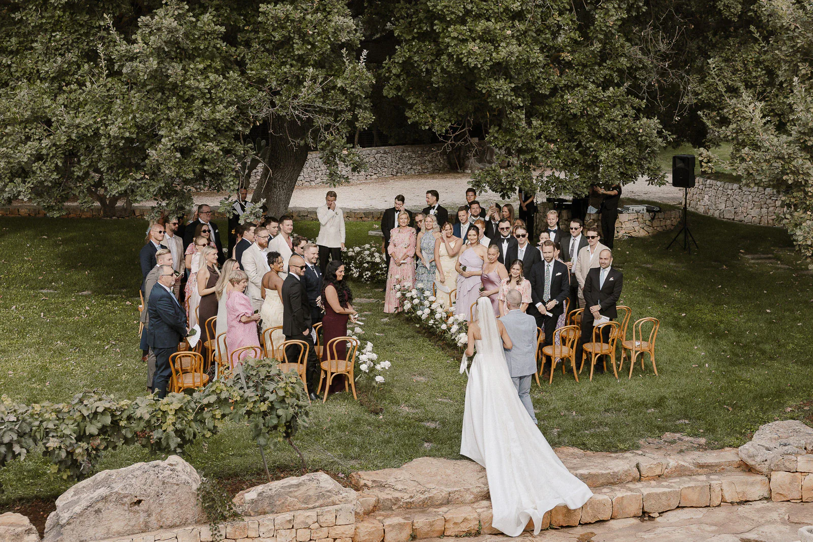 In Puglia, guests watch as the bride approaches her groom beneath ancient trees, an intimate ceremony unfolding amid tranquil greenery.