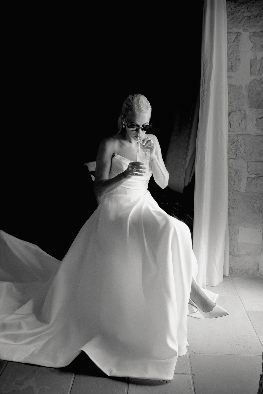A bride in a strapless gown savors a quiet moment by the window in sunlit Puglia, elegant sunglasses completing her look.