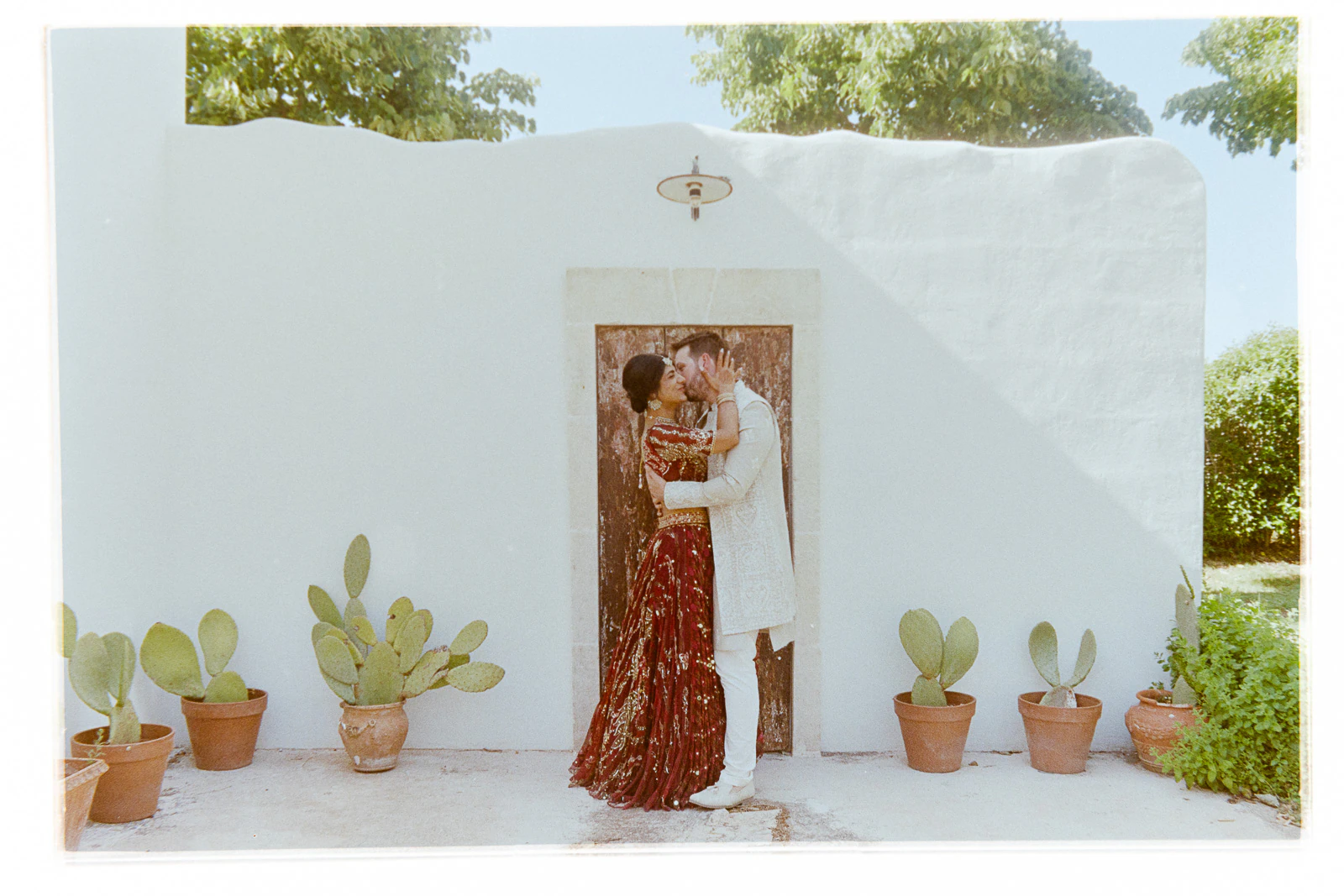 An elegant couple shares a kiss before a rustic wooden door in Puglia, framed by white stucco and lush Mediterranean greenery.