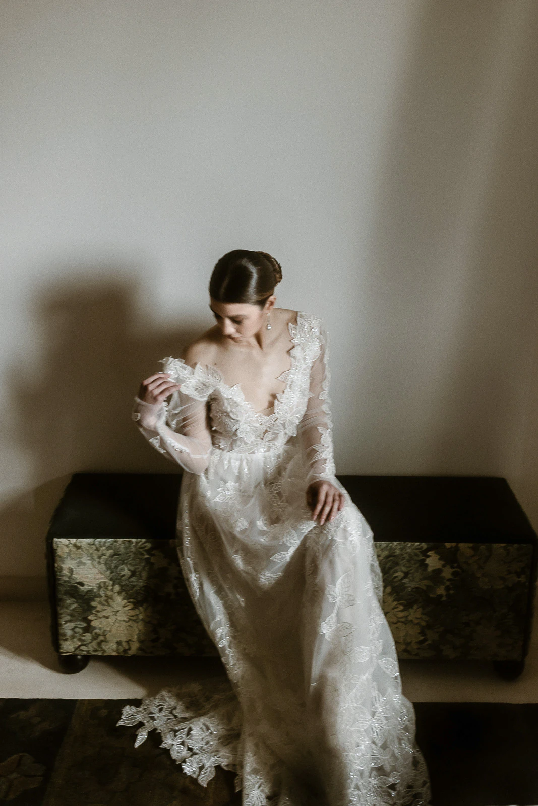 In Puglia, a bride in an elegant lace gown sits quietly on a patterned bench, lost in intimate reflection.