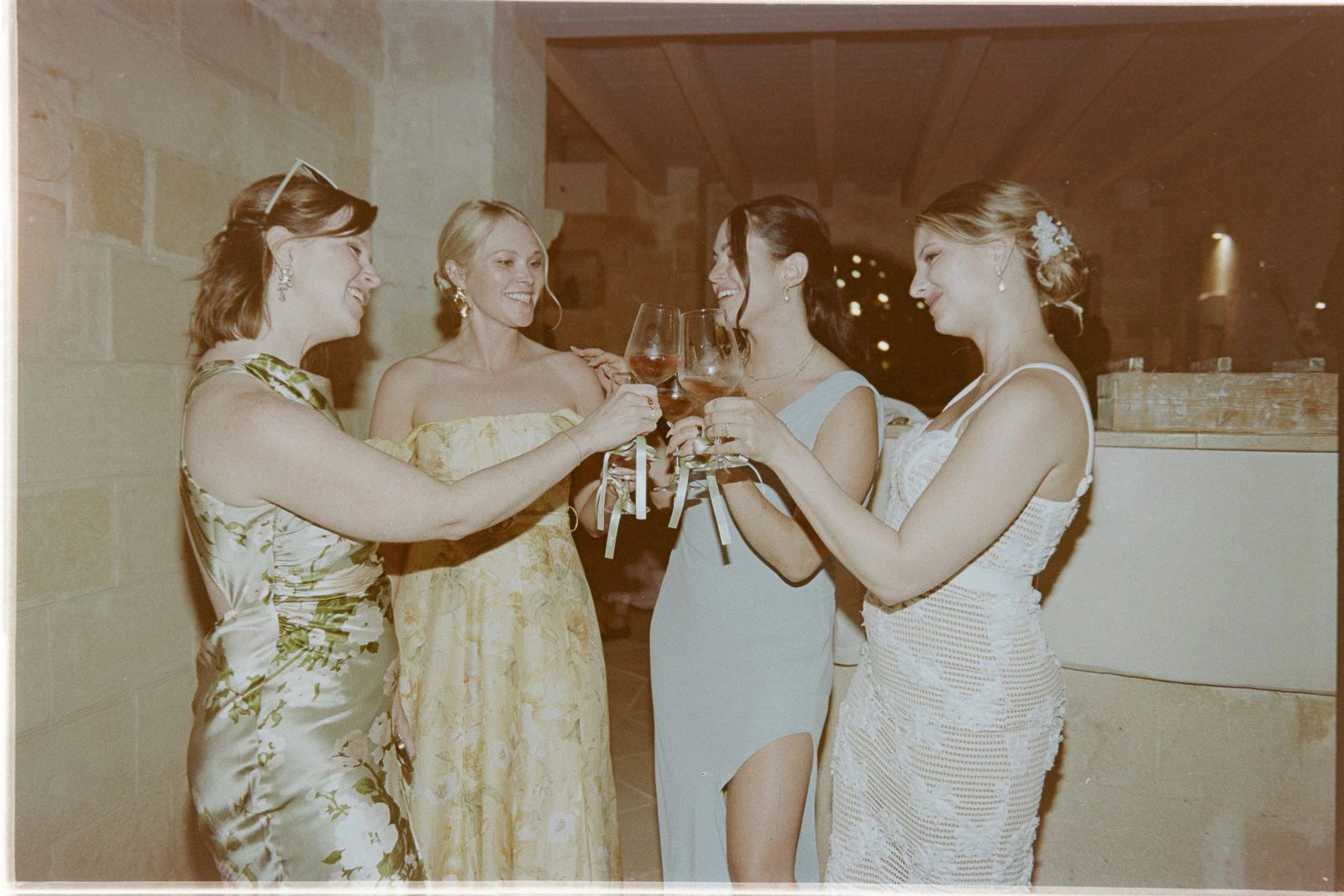 In a sunlit Italian villa, four elegantly dressed women share laughter and toast with wine, capturing joyful wedding moments.