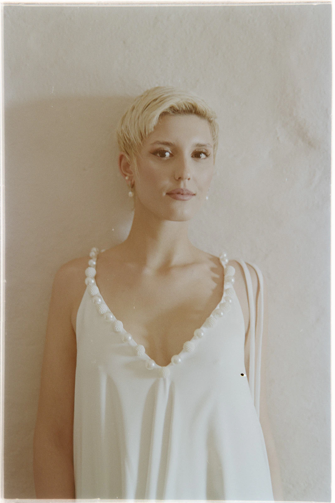 With short blonde hair, she radiates elegance in a pearl-detailed white gown, poised against sunlit Italian walls.