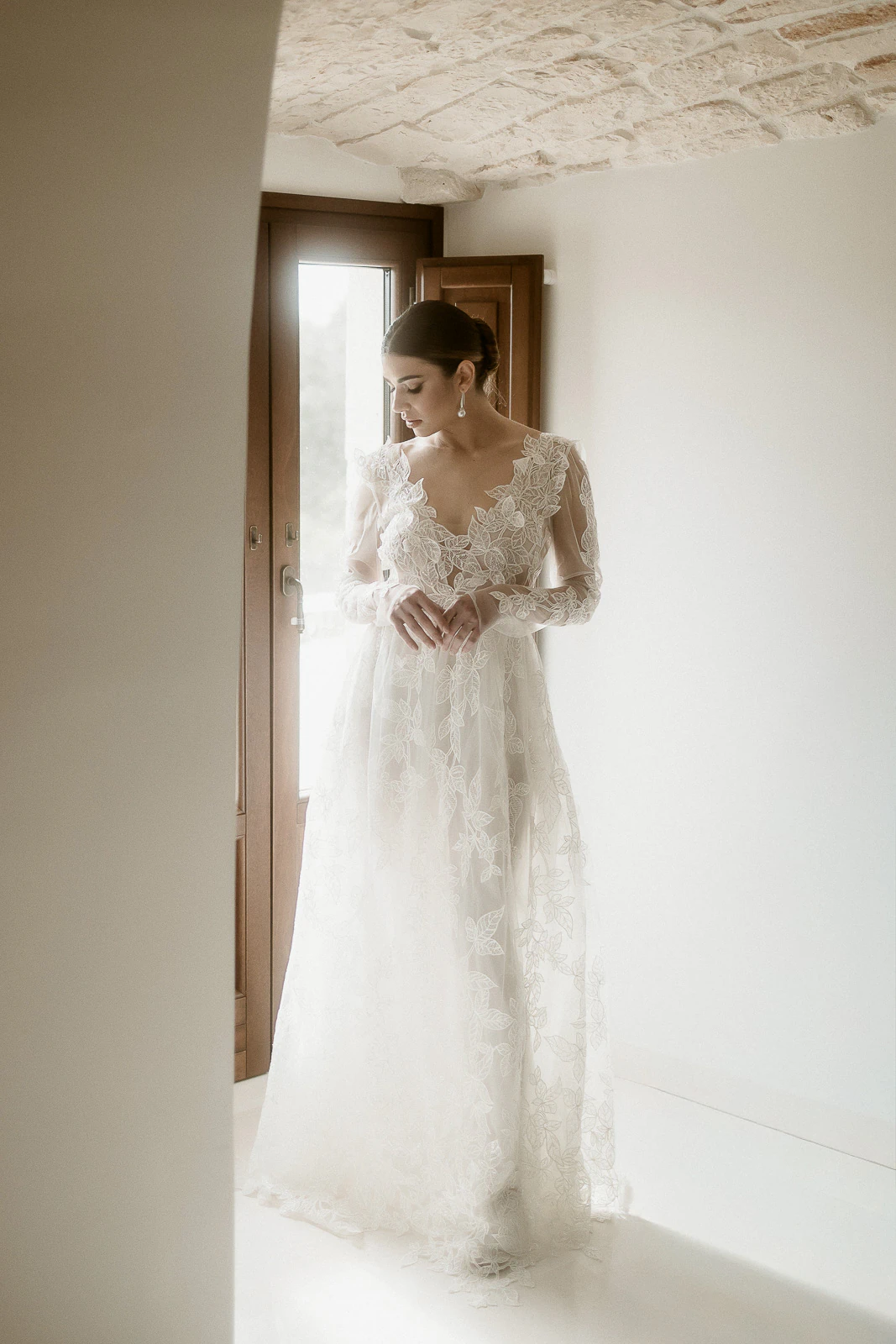 A bride in delicate lace gazes downward by a sunlit window, serene moments before her elegant destination wedding in Puglia, Italy.