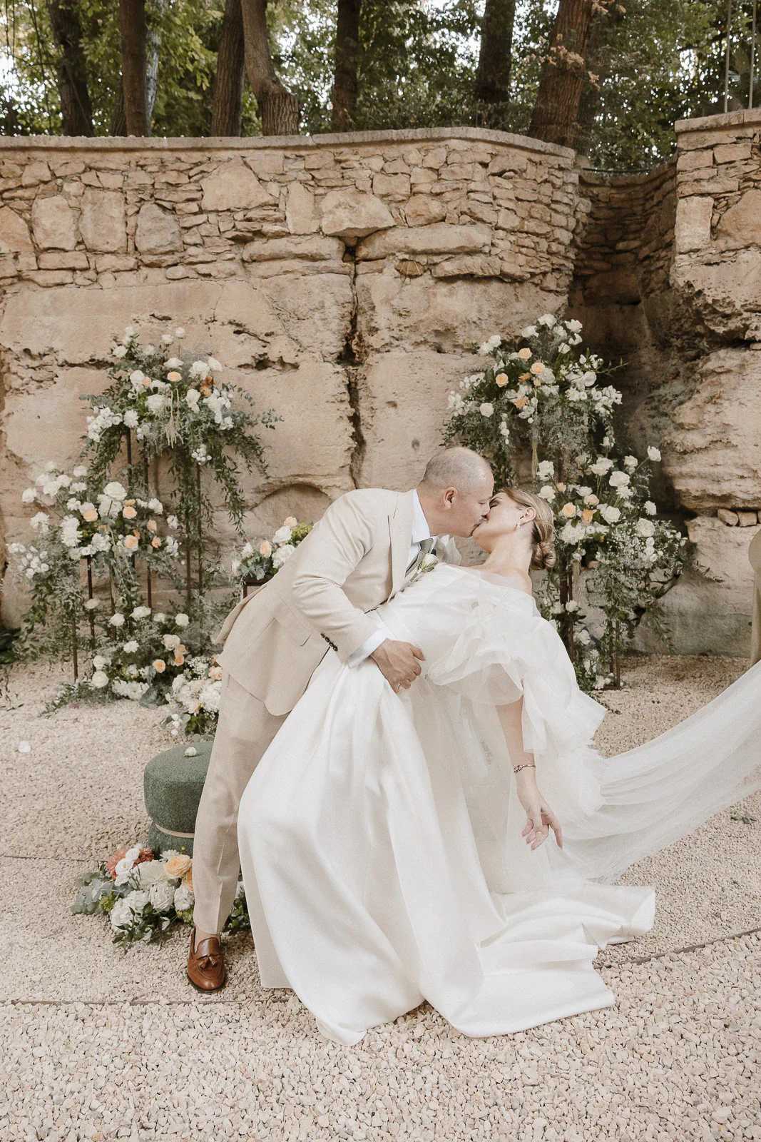 A groom in a beige suit dips and kisses his bride amid sunlit stone walls and lush florals at a romantic wedding in Puglia.