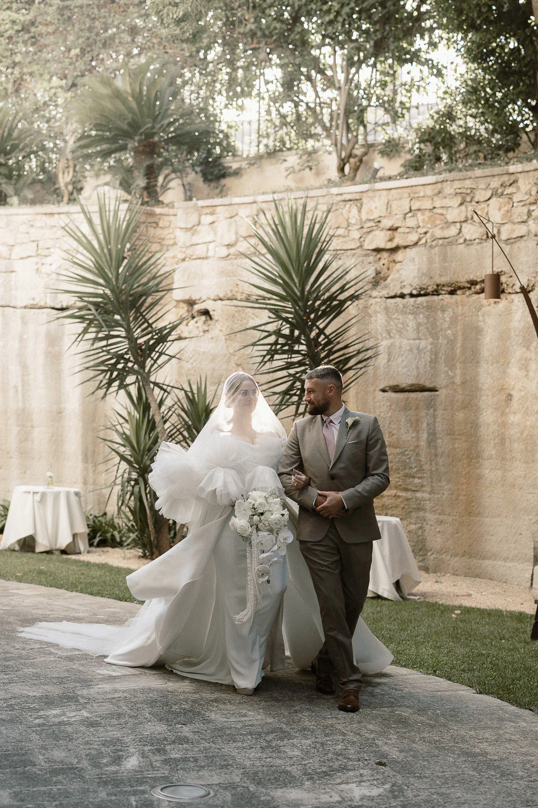 A bride and her escort walk arm-in-arm beside ancient stone walls, embraced by lush greenery under the Puglian sun.