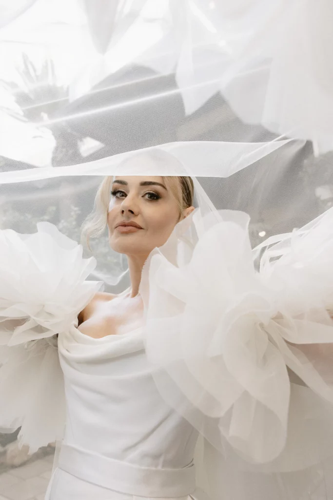 Beneath a sheer veil, a bride in voluminous tulle sleeves meets the camera’s gaze in sun-drenched Puglia, Italy.