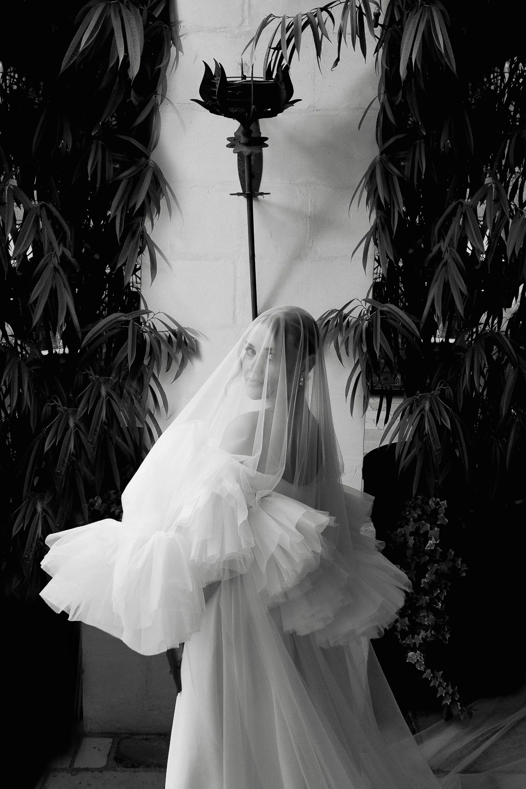 A bride glances back amid sunlit greenery and whitewashed Puglian walls, her gown and veil catching the soft Italian light.