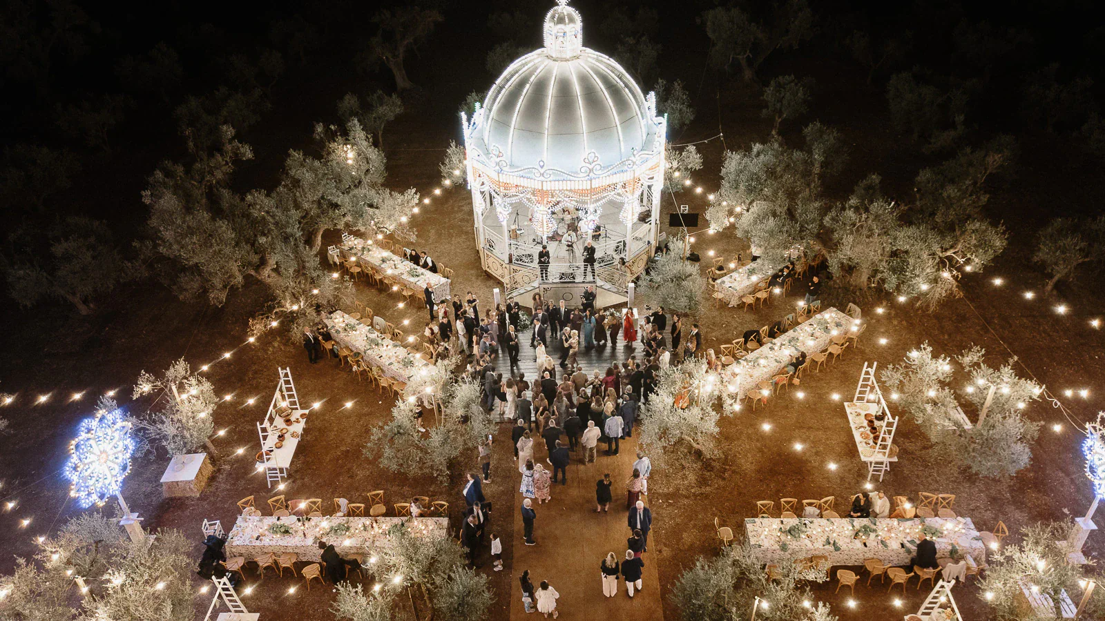 A luminous gazebo anchors a romantic reception under twinkling lights, as guests dine beneath ancient trees in Puglia, Italy.