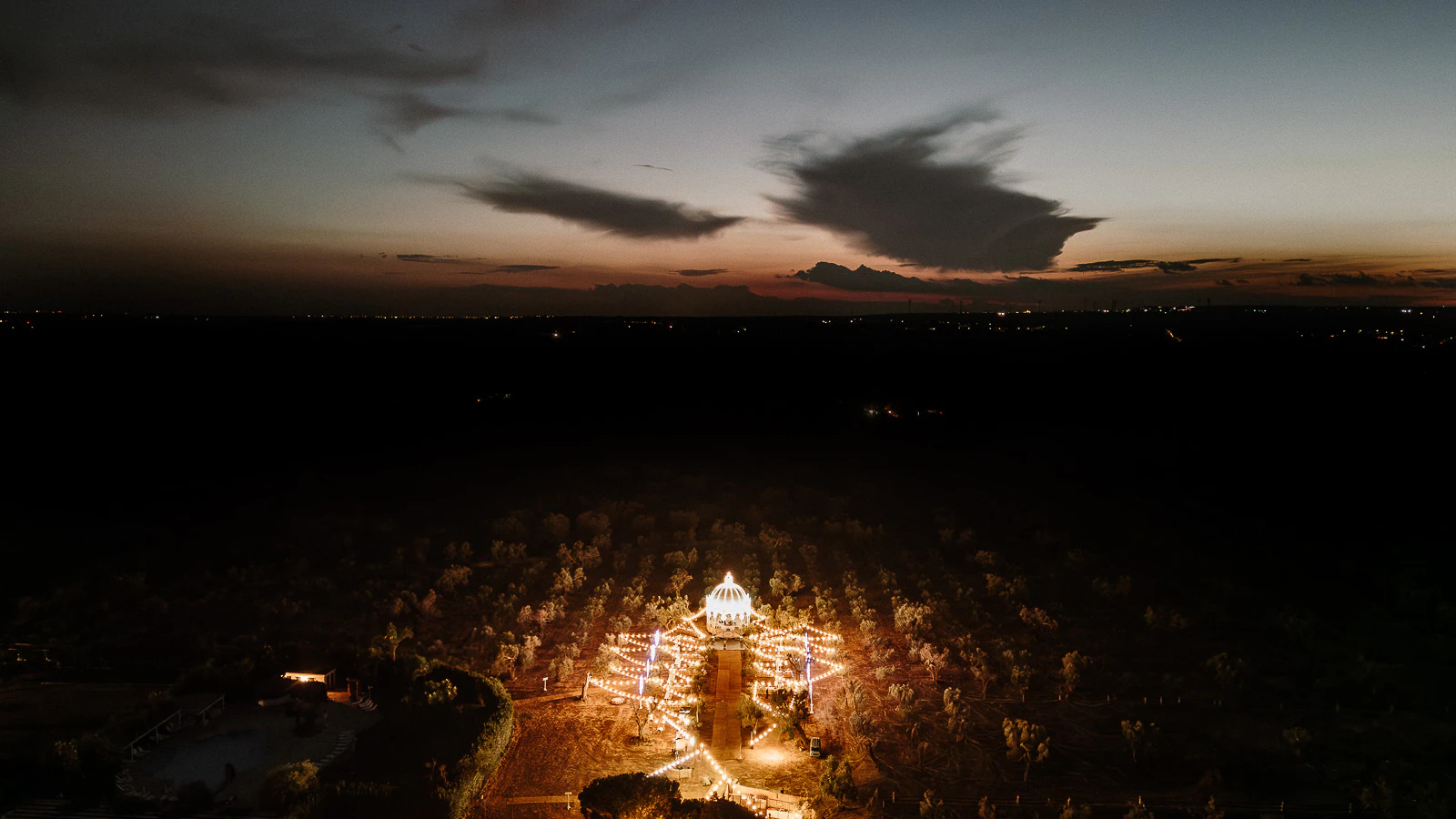A dusk aerial of a Puglia villa’s dome aglow with string lights, cradled by olive trees beneath a cinematic Italian sky.