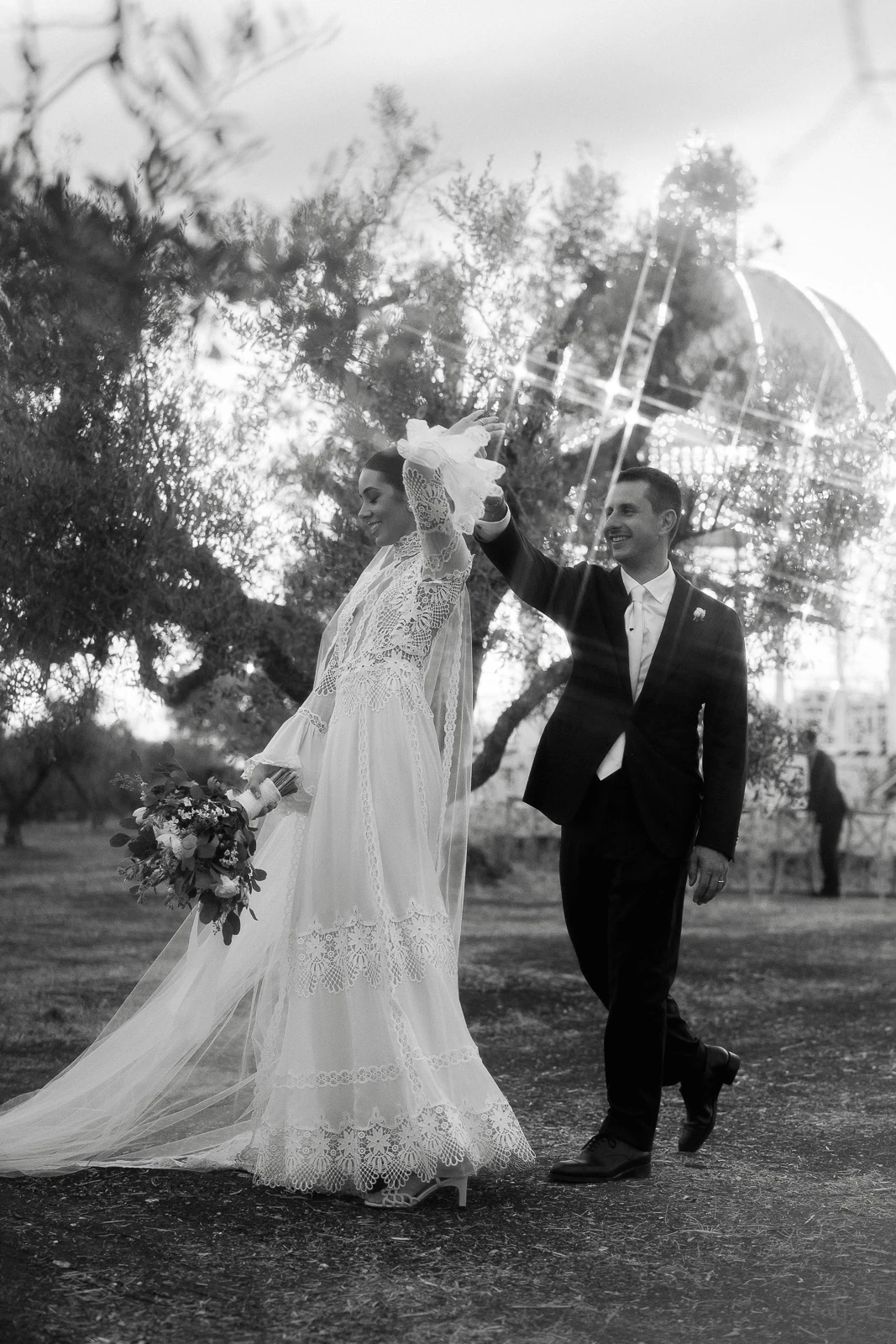 Amid olive groves in Puglia, a groom gently leads his bride in lace and veil through sunlit Italian countryside.