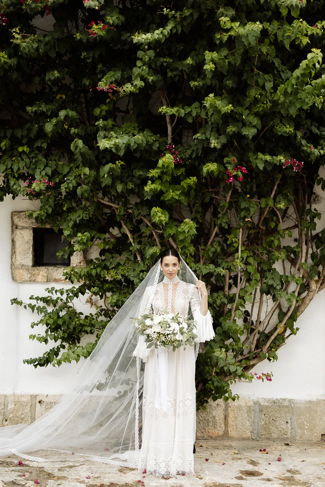 An elegant bride in a flowing white gown poses by lush vines against whitewashed walls, capturing Puglia’s romantic essence.