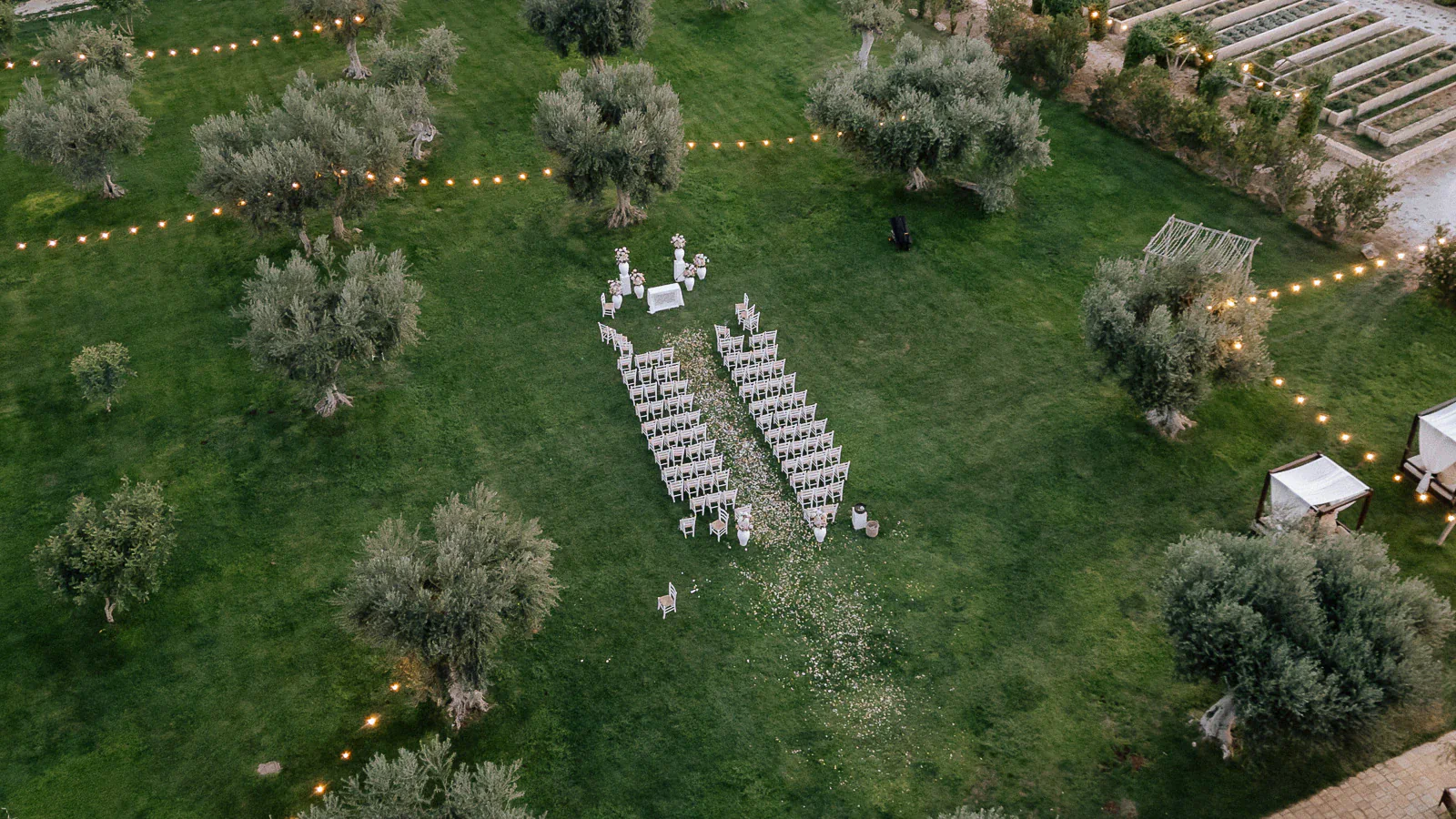 Aerial scene of an intimate Puglia wedding ceremony, elegant chairs on verdant lawn beneath twinkling string lights and ancient trees.